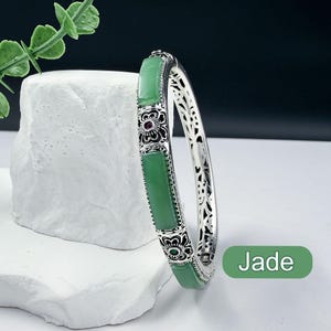 May include: A silver bangle bracelet with rectangular green jade stones and ornate floral accents. The bracelet has a hinged design and is displayed on a white textured surface. The word "Jade" is in a green box.