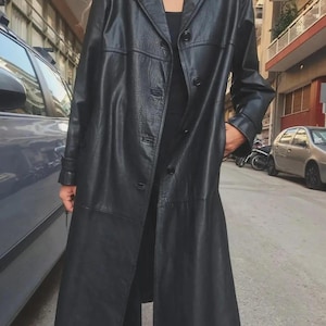 May include: A long black leather coat featuring a classic collar, a button-down front closure, and long sleeves. The coat has a smooth texture with visible seams across the chest and waist, providing a structured fit that extends to the mid-calf.
