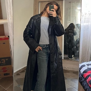 May include: A long black leather trench coat with a button-down front, wide lapels, and long sleeves, worn over a light gray crew-neck t-shirt and dark blue denim jeans. The coat features a structured collar and visible buttons down the center.