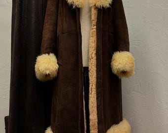 May include: A long, dark brown coat featuring a thick, cream-colored faux fur trim along the large collar, the sleeve cuffs, and the bottom hem. The coat is displayed on a mannequin and has a visible cream-colored faux fur lining throughout.