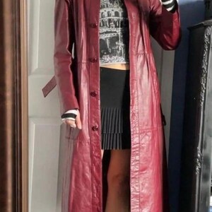 May include: A long, deep red leather trench coat with a button-front closure and a collar, worn over a black and white graphic t-shirt, a black pleated mini skirt, and black platform ankle boots with a block heel and striped long sleeves.