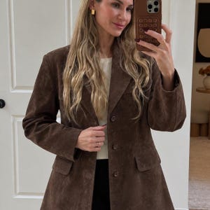 Pode incluir: Um blazer de camurça castanho-escuro com lapelas entalhadas, mangas compridas e fecho frontal de três botões. O casaco inclui dois bolsos com aba na cintura e é usado sobre uma blusa branca canelada. O material de camurça tem textura mate.
