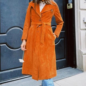 May include: A long, burnt orange suede coat featuring a wide notched collar, a button-front closure, and a matching tie belt at the waist. The coat has long sleeves, two side pockets, and a structured silhouette that falls below the knee.