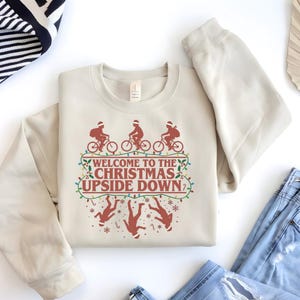 May include: Beige sweatshirt with a graphic featuring three figures on bicycles wearing Santa hats, the text "WELCOME TO THE CHRISTMAS UPSIDE DOWN," and upside-down figures. The design is in shades of brown and red.