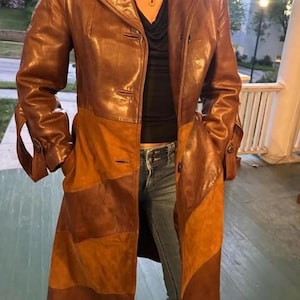 May include: A long, multi-textured coat featuring a combination of smooth brown leather and panels of tan suede. The coat has a button-front closure, long sleeves with cuff details, and a structured collar, worn over a black top and blue denim jeans.