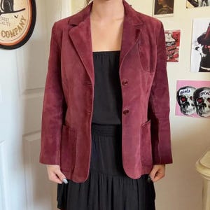 May include: A deep burgundy suede blazer featuring a notched lapel, two front buttons, two large patch pockets at the waist, and a single chest pocket. The jacket is worn over a black top and skirt, showcasing a structured fit and long sleeves.