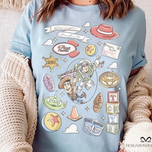 May include: Light blue t-shirt featuring a colorful graphic design with characters and elements from the animated film series. The design includes the text "Son is got a Friend in me", "ANDY'S ROOM KEEP OUT", "SHERIFF", and "ANDY".