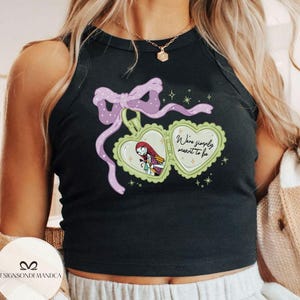 Puede incluir: Camiseta sin mangas negra con un dise&ntilde;o de medall&oacute;n en forma de coraz&oacute;n, con un personaje de dibujos animados y el texto "We're simply meant to be". El medall&oacute;n est&aacute; adornado con un lazo morado y estrellas. La camiseta es sin mangas.