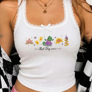 May include: A white ribbed tank top with lace trim along the neckline and a small center bow. The front features a graphic print of a green chameleon, a golden flower, a crown, and lanterns with the text Best Day ever in black script.