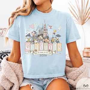 Pu&ograve; includere: T-shirt azzurra con una grafica colorata con personaggi Disney, un castello e la parola "Graduate" in colori pastello. &Egrave; incluso anche il testo "From the Tassel to the Castle 2026". La maglietta &egrave; a girocollo.