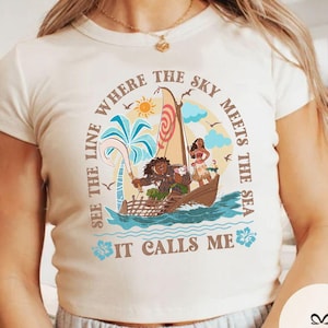 May include: Cream-colored t-shirt featuring a colorful illustration of a boat with characters, palm trees, and the sun. The text on the shirt reads "Where the sky meets the sea, see the line, it calls me."