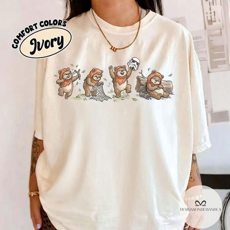 K&ouml;nnte beinhalten: Elfenbeinfarbenes Comfort Colors T-Shirt mit einer verspielten Ewok-Illustration. Das Design zeigt Ewoks mit einem Netz, einem Stormtrooper-Helm und einem Baumstamm. Das Shirt tr&auml;gt die Aufschrift "Comfort Colors" und "Ivory".