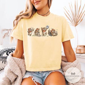 K&ouml;nnte beinhalten: Hellgelbes T-Shirt mit einer Cartoon-Illustration von Ewoks in verschiedenen Posen, darunter eine, die einen Stormtrooper-Helm h&auml;lt. Das Design ist auf einem kurz&auml;rmeligen T-Shirt abgebildet. Das Shirt hat eine lockere Passform und ist aus weichem Material.