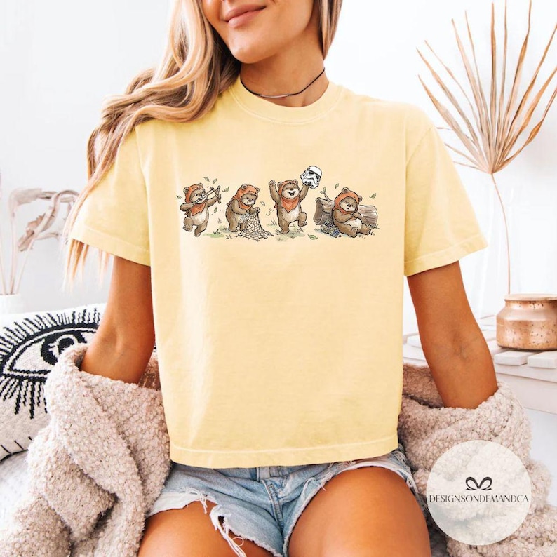 K&ouml;nnte beinhalten: Hellgelbes T-Shirt mit einer Cartoon-Illustration von Ewoks in verschiedenen Posen, darunter eine, die einen Stormtrooper-Helm h&auml;lt. Das Design ist auf einem kurz&auml;rmeligen T-Shirt abgebildet. Das Shirt hat eine lockere Passform und ist aus weichem Material.