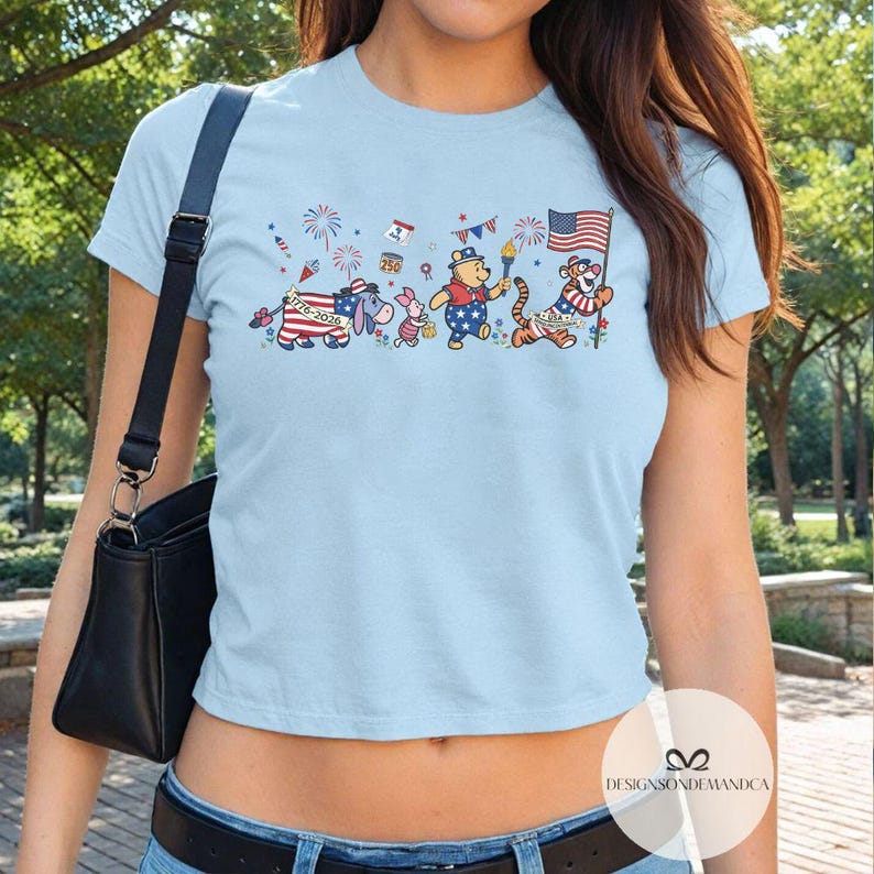 May include: Light blue t-shirt featuring a patriotic design with cartoon characters. The design includes Winnie the Pooh, Piglet, Eeyore, and Tigger, each adorned with American flag-themed attire, celebrating the 4th of July. The text "1776-2020" is also visible.