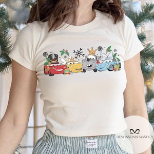 May include: Cream-colored t-shirt featuring a festive graphic of animated cars adorned with holiday decorations. The cars are in various colors, including red, yellow, gray, and blue, with crowns, bows, and snowflakes. The shirt is a short-sleeved crew neck.