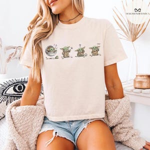 May include: Cream-colored t-shirt featuring a graphic of a character from a popular science fiction series. The graphic shows the character in four different poses. The word "cool" is printed on the shirt. The shirt is paired with denim shorts.