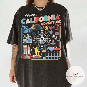 以下が含まれることがあります： ディズニー・カリフォルニア・アドベンチャーのカラフルなグラフィックが特徴の濃いグレーのTシャツ。ミッキーマウス、カーズのキャラクター、その他のパークのアイコンが描かれた観覧車がデザインされています。「Disney California Adventure」の文字が上部にプリントされています。