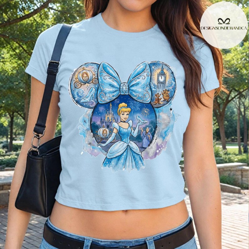 Puede incluir: Camiseta azul claro con un dise&ntilde;o tem&aacute;tico de Cenicienta. El dise&ntilde;o incluye un gran lazo azul, Cenicienta, un carruaje y un castillo. La camiseta tiene mangas cortas y cuello redondo. El texto "DESIGNSONDEMANDCA" es visible en la parte superior derecha.