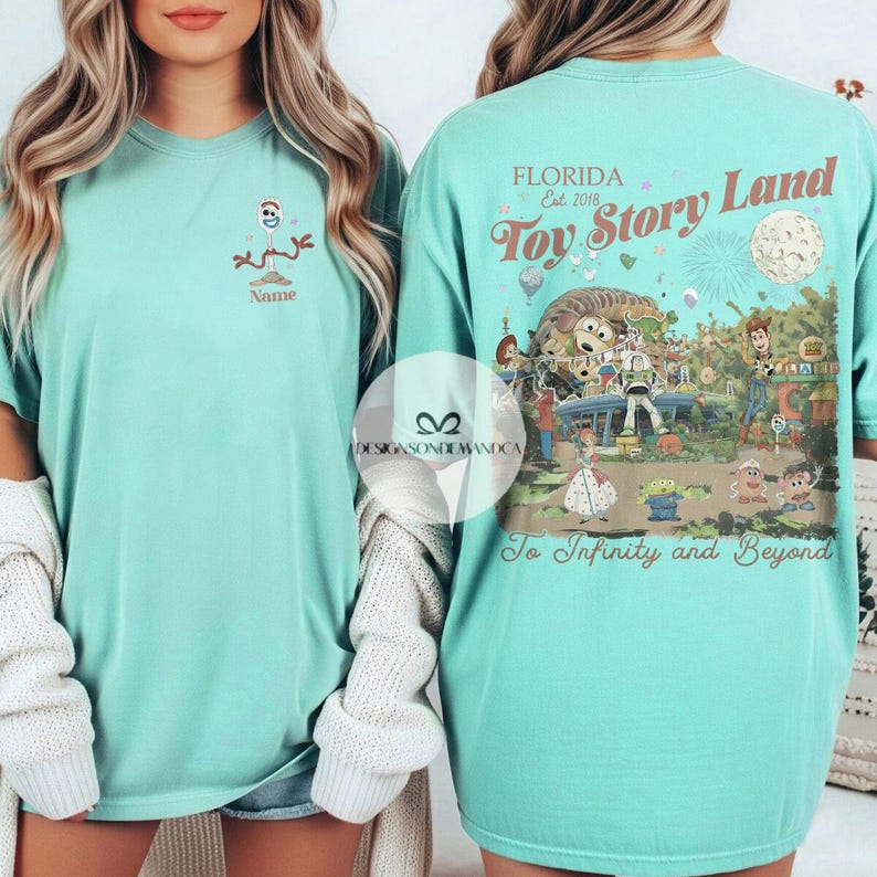 Puede incluir: Una camiseta de color verde azulado claro con un dise&ntilde;o de dibujos animados de Forky y la palabra "Name" debajo. La parte trasera de la camiseta muestra un colorido dise&ntilde;o de "Toy Story Land" con el texto "Florida Est. 2018" y "To Infinity and Beyond."