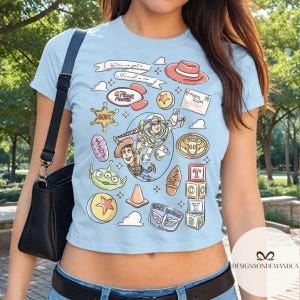 May include: Light blue crop top featuring colorful cartoon graphics of characters and elements from the movie Toy Story. The design includes Woody, Buzz Lightyear, and other iconic imagery, with text elements like "Andy's Room" and "Sheriff".