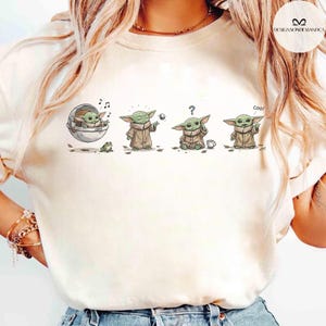 May include: Cream-colored t-shirt featuring a cartoon illustration of a character in various poses. The character is depicted in a pod, holding a ball, with a question mark, and giving a thumbs up with the word "Cool!"