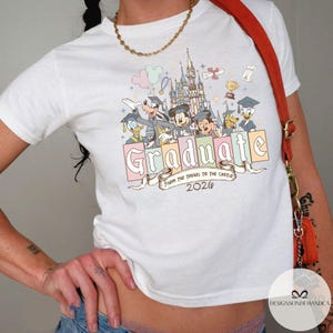 Pu&ograve; includere: T-shirt bianca con personaggi Disney come Topolino, Paperino e Pippo, una grafica del castello e la parola "Graduate" in colori pastello. &Egrave; presente anche il testo "From the Tassel to the Castle 2020".