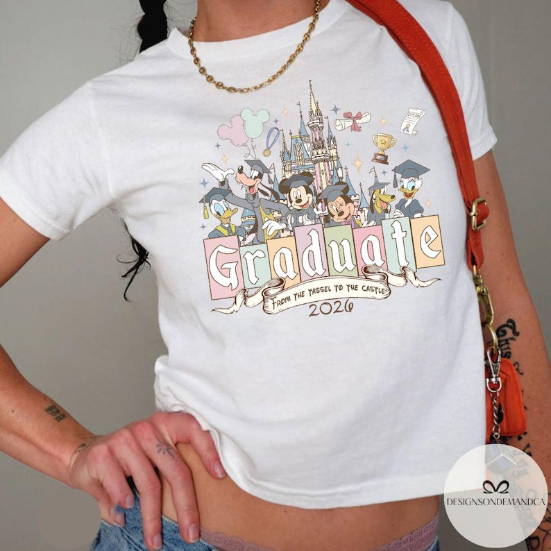 Pu&ograve; includere: T-shirt bianca con personaggi Disney come Topolino, Paperino e Pippo, una grafica del castello e la parola "Graduate" in colori pastello. &Egrave; presente anche il testo "From the Tassel to the Castle 2020".