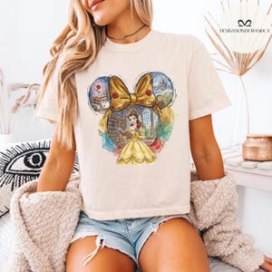 Puede incluir: Camiseta color crema con un gr&aacute;fico colorido de Bella de La Bella y la Bestia dentro de un contorno de oreja de Mickey Mouse. El gr&aacute;fico incluye un lazo amarillo, una rosa y un castillo. La camiseta es de estilo casual.