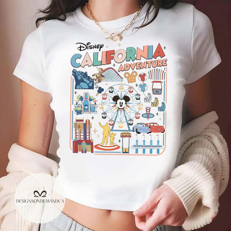 以下が含まれることがあります： 「Disney California Adventure」の文字とカラフルなグラフィックデザインが施された白いTシャツ。ミッキーマウスの観覧車、ジェットコースター、その他のパークアトラクションがデザインされています。半袖のシャツです。