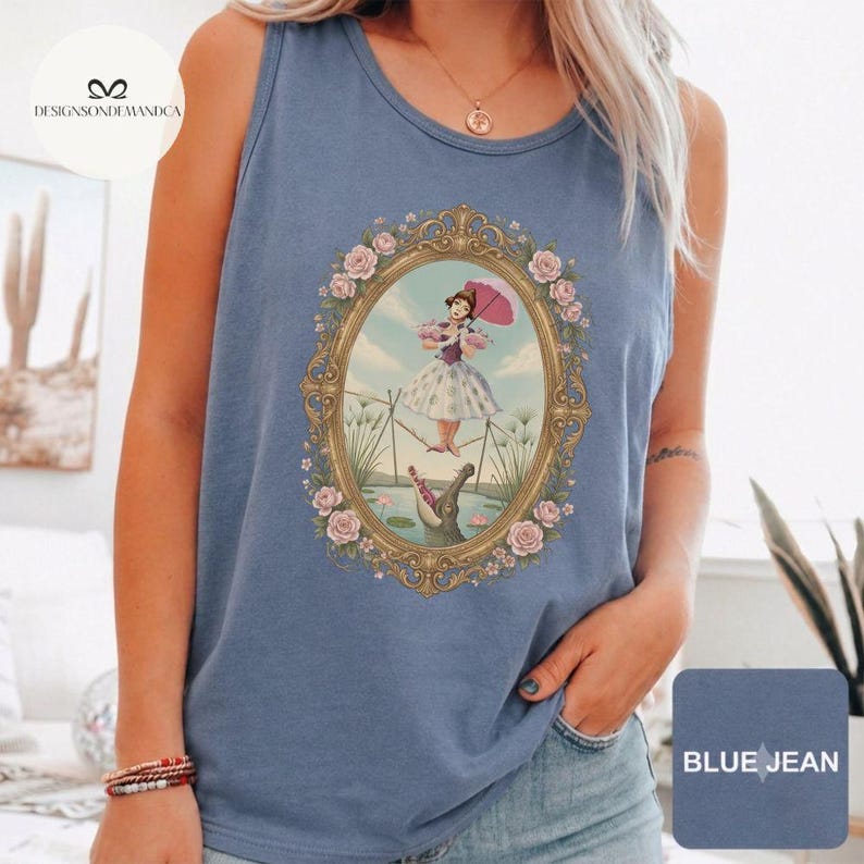 May include: A blue jean colored tank top featuring a central graphic of a person in a white dress holding a pink umbrella, balancing on a tightrope above a crocodile, all enclosed within an ornate gold oval frame decorated with pink roses.
