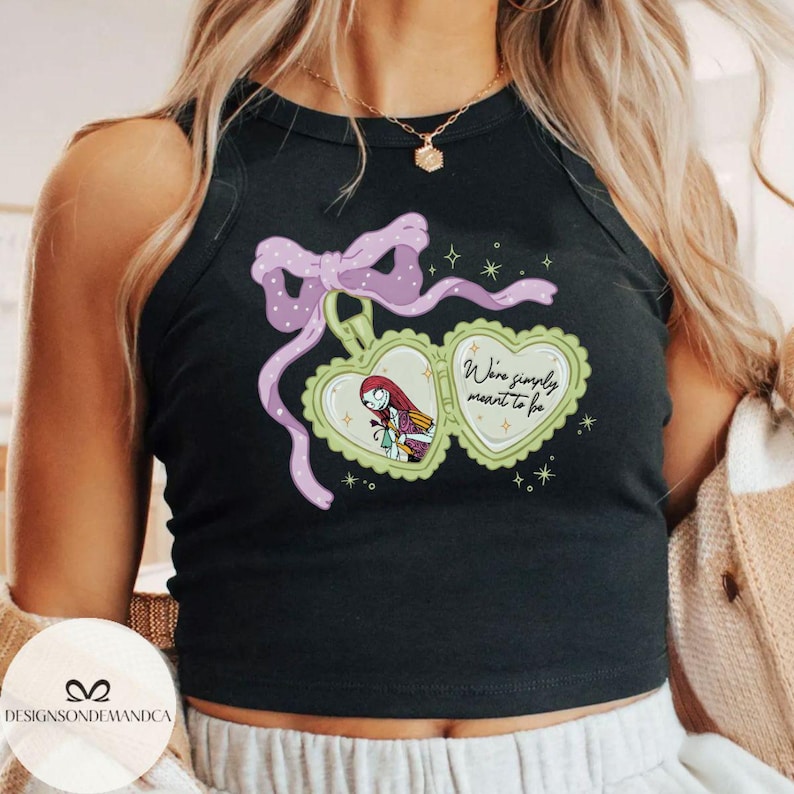 Puede incluir: Camiseta sin mangas negra con un dise&ntilde;o de medall&oacute;n en forma de coraz&oacute;n, con un personaje de dibujos animados y el texto "We're simply meant to be". El medall&oacute;n est&aacute; adornado con un lazo morado y estrellas. La camiseta es sin mangas.