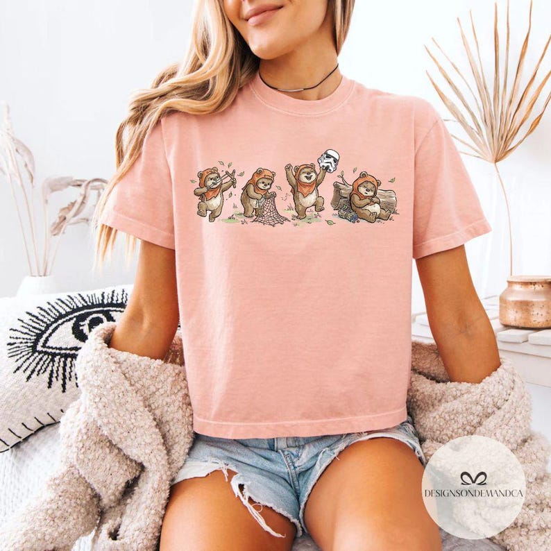 K&ouml;nnte beinhalten: Hellrosa T-Shirt mit einer Cartoon-Illustration von Ewoks. Die Ewoks tragen braune Kapuzenoutfits und interagieren mit einem Stormtrooper-Helm und einem Baumstamm. Das T-Shirt hat einen Rundhalsausschnitt und kurze &Auml;rmel.