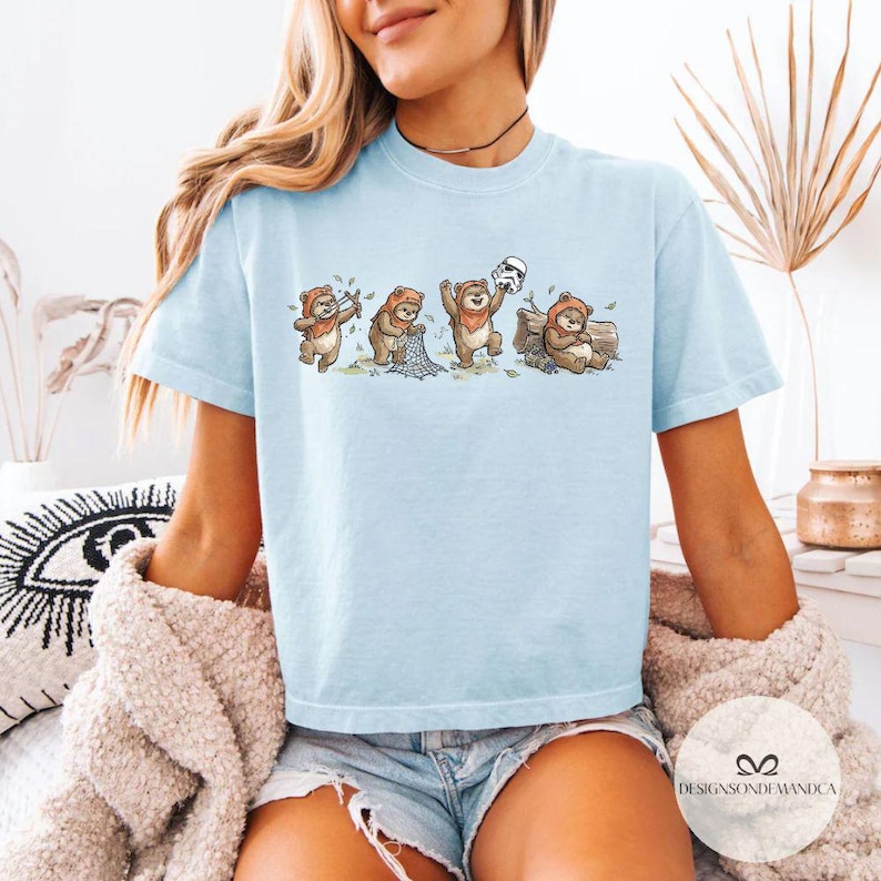 K&ouml;nnte beinhalten: Hellblaues T-Shirt mit einer skurrilen Illustration von Ewoks. Die Cartoon-Ewoks werden mit Pfeil und Bogen, einem Netz, einem Stormtrooper-Helm und auf einem Baumstamm sitzend dargestellt. Das Shirt hat den Text "DESIGNSONDEMANDCA".