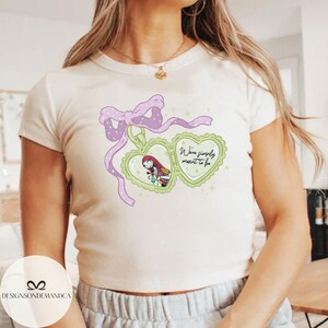 Puede incluir: Camiseta color crema con un colgante en forma de coraz&oacute;n con un personaje de dibujos animados y el texto "We're simply meant to be". El colgante est&aacute; adornado con un lazo y una cinta morados.
