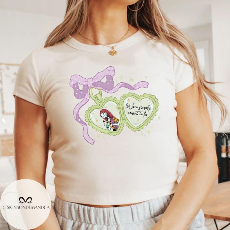Puede incluir: Camiseta color crema con un colgante en forma de coraz&oacute;n con un personaje de dibujos animados y el texto "We're simply meant to be". El colgante est&aacute; adornado con un lazo y una cinta morados.