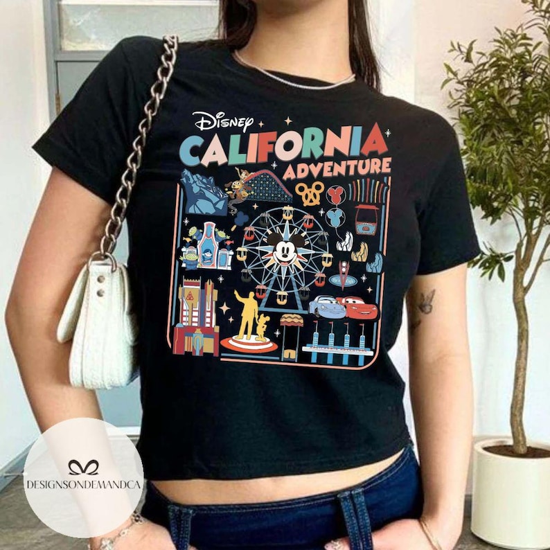 以下が含まれることがあります： ディズニー・カリフォルニア・アドベンチャーのテーマパークのカラフルなグラフィックが特徴の黒いTシャツ。ミッキーマウス、カーズのキャラクター、その他のパークのアイコンがデザインされています。「Disney California Adventure」の文字が上部に表示されています。