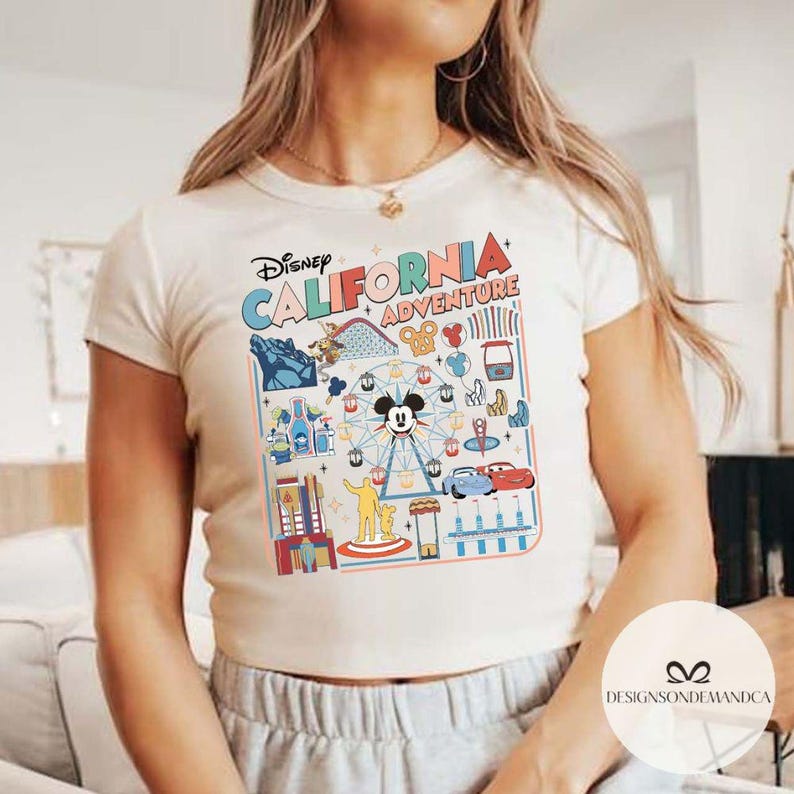 以下が含まれることがあります： ディズニー・カリフォルニア・アドベンチャーのカラフルなグラフィックが特徴のクリーム色のTシャツ。デザインには、観覧車に乗ったミッキーマウス、ジェットコースター、その他のパークアイコンが含まれています。「Disney California Adventure」の文字が上部にアーチ状に配置されています。