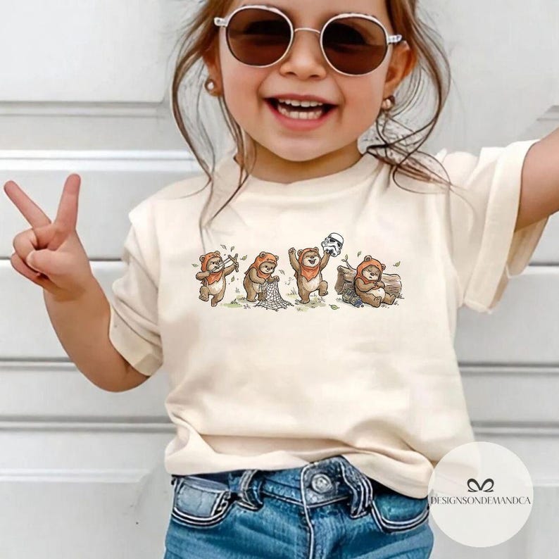K&ouml;nnte beinhalten: Cremefarbenes T-Shirt mit einer Grafik von Ewoks in verschiedenen Posen. Die Cartoon-Ewoks sind mit B&ouml;gen, Netzen und einem Stormtrooper-Helm dargestellt. Das Shirt wird von einem Kind getragen, das eine Sonnenbrille und blaue Jeans tr&auml;gt.