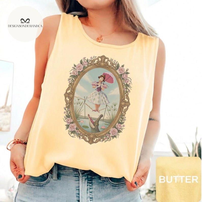May include: A pale yellow sleeveless tank top featuring a central graphic print of a person in a dress holding a pink umbrella, walking a tightrope above a crocodile, all enclosed within an ornate gold-toned oval frame decorated with pink roses.