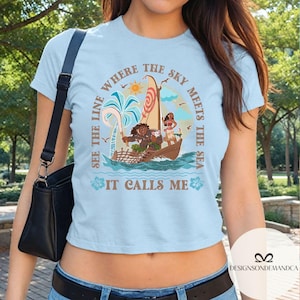 May include: Light blue t-shirt featuring a nautical scene with the text "SEE THE LINE WHERE THE SKY MEETS THE SEA IT CALLS ME." The design includes a boat with characters, palm trees, and a sun.