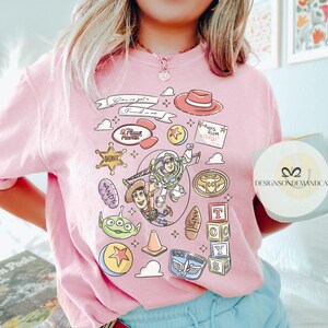 May include: Pink t-shirt featuring a colorful graphic design with characters and elements from the animated film Toy Story. The design includes Woody, Buzz Lightyear, and other iconic imagery. The text "You've got a friend in me" is also visible.