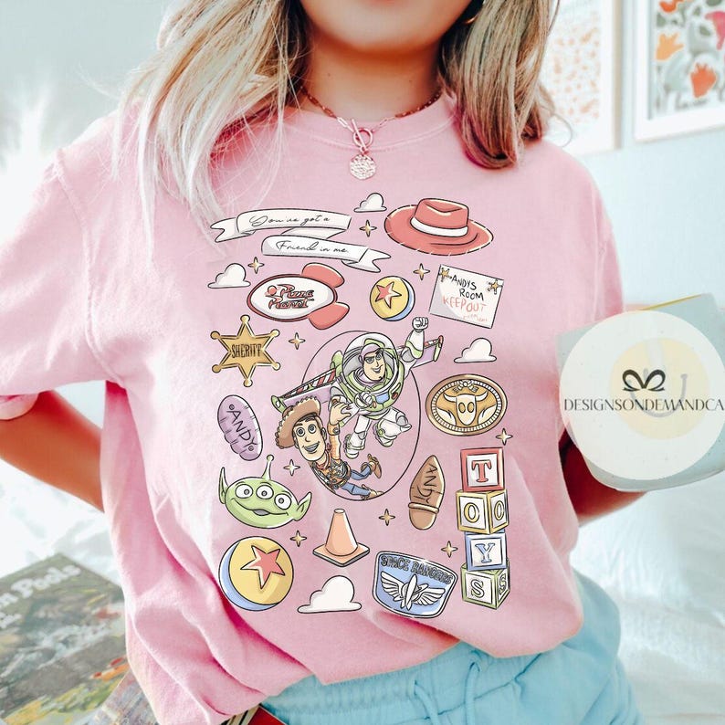 May include: Pink t-shirt featuring a colorful graphic design with characters and elements from the animated film Toy Story. The design includes Woody, Buzz Lightyear, and other iconic imagery. The text "You've got a friend in me" is also visible.