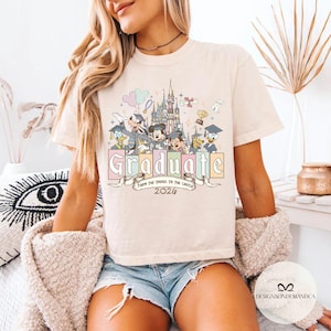 Pu&ograve; includere: T-shirt color crema con una grafica colorata di personaggi Disney, un castello e la parola "Graduate" in colori pastello. &Egrave; incluso anche il testo "From the Tassel to the Castle 2026". La maglietta &egrave; in stile casual.