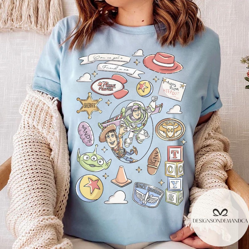May include: Light blue t-shirt featuring a colorful graphic design with characters and elements from the animated film series. The design includes the text "Son is got a Friend in me", "ANDY'S ROOM KEEP OUT", "SHERIFF", and "ANDY".