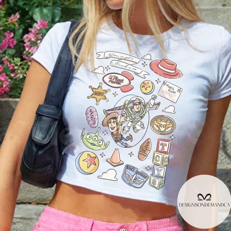 May include: White crop top featuring a colorful graphic design with characters and elements from the animated film series. The design includes Woody, Buzz Lightyear, and other iconic imagery. The text "You've got a friend in me" is also included.