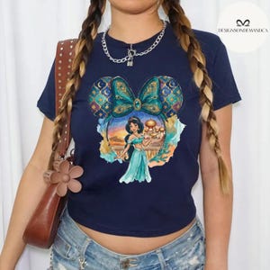 Puede incluir: Camiseta azul marino con un estampado colorido de la Princesa Jasmine frente a un palacio, enmarcada por orejas de Mickey Mouse con dise&ntilde;os de plumas de pavo real. La camiseta tiene cuello redondo y mangas cortas.