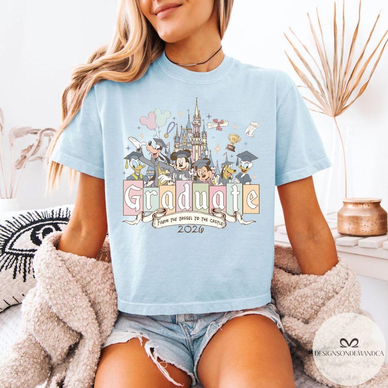 Pu&ograve; includere: T-shirt azzurra con una grafica colorata con personaggi Disney, un castello e la parola "Graduate" in colori pastello. &Egrave; incluso anche il testo "From the Tassel to the Castle 2026". La maglietta &egrave; a girocollo.