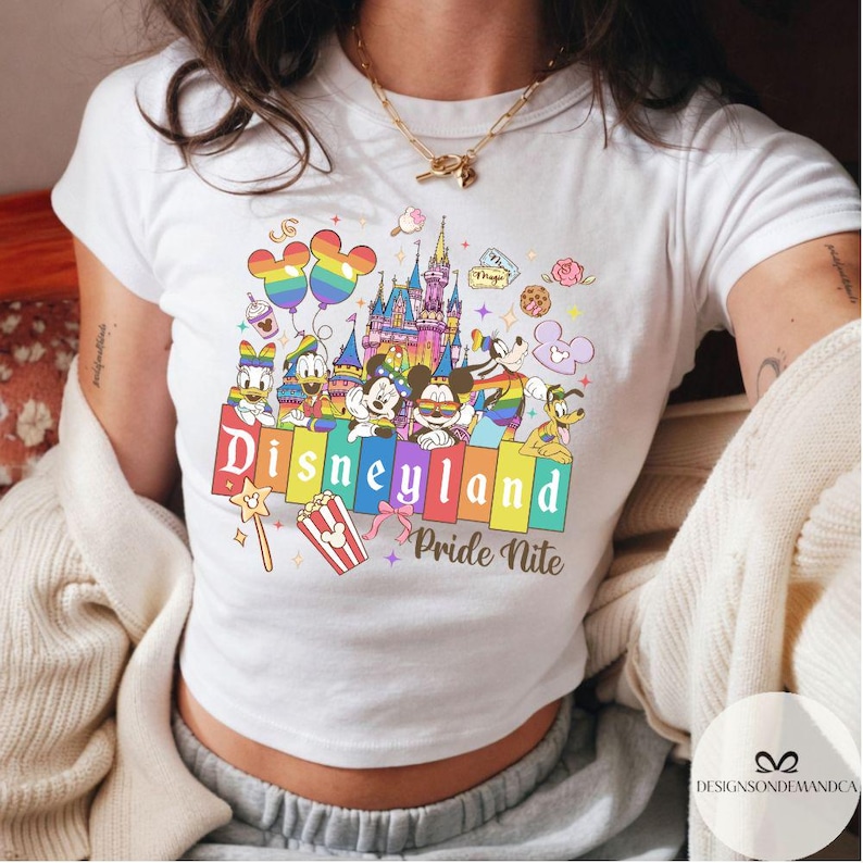May include: White t-shirt with a vibrant Disneyland Pride Nite design. The graphic showcases Disney characters, a castle, balloons, and the text "Disneyland Pride Nite" in rainbow colours. A popcorn box and a magic wand are also included in the design.