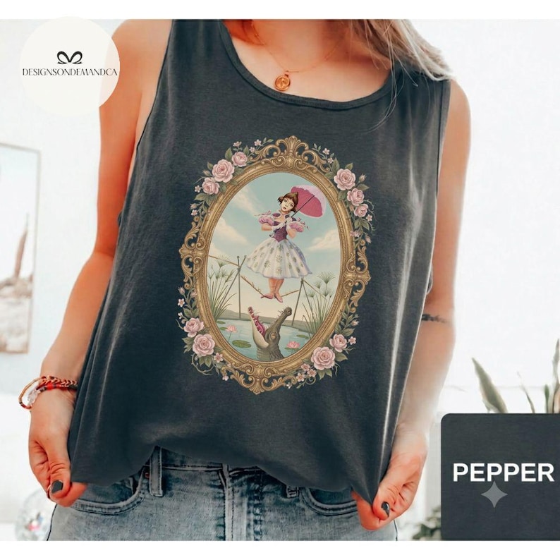 May include: A charcoal gray sleeveless tank top featuring a central graphic print of a person in a white dress holding a pink umbrella, walking a tightrope above an alligator, all enclosed within an ornate gold oval frame with pink roses.